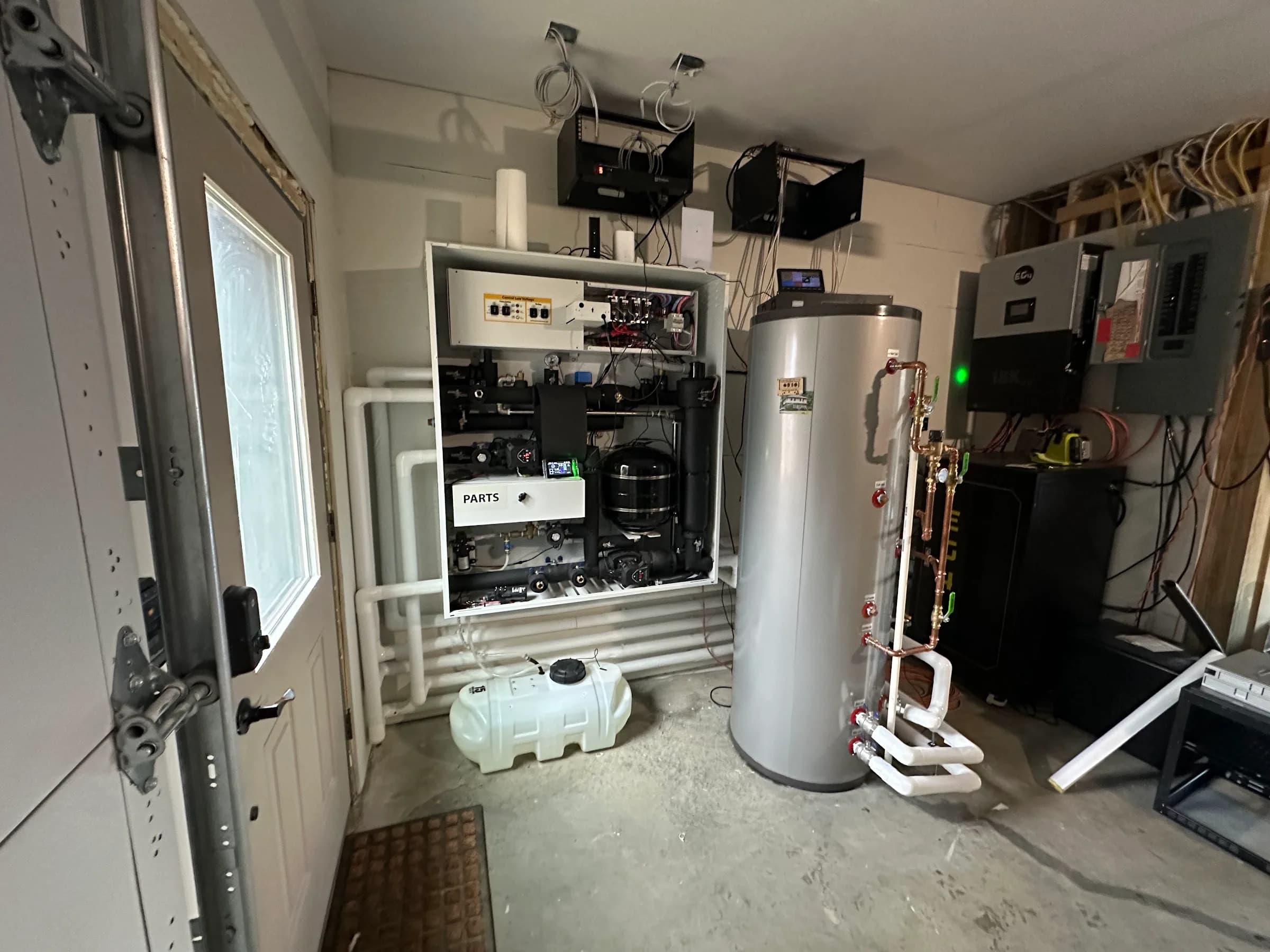 An air to water heat pump panel and buffer tank installed along a garage wall with the overhead door open