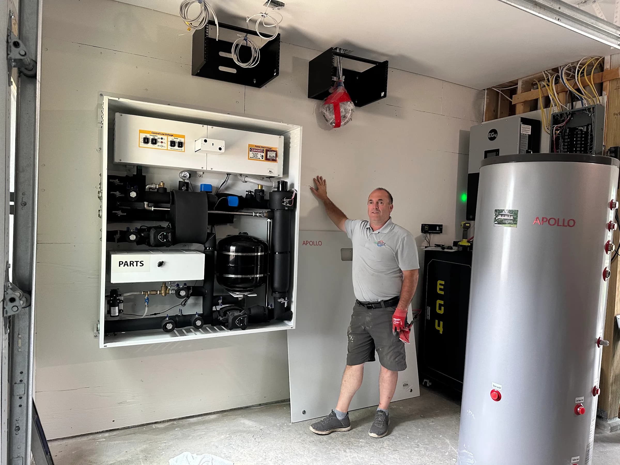 A technician standing beside a wall mounted air to water heat pump panel and a tall buffer tank in a garage