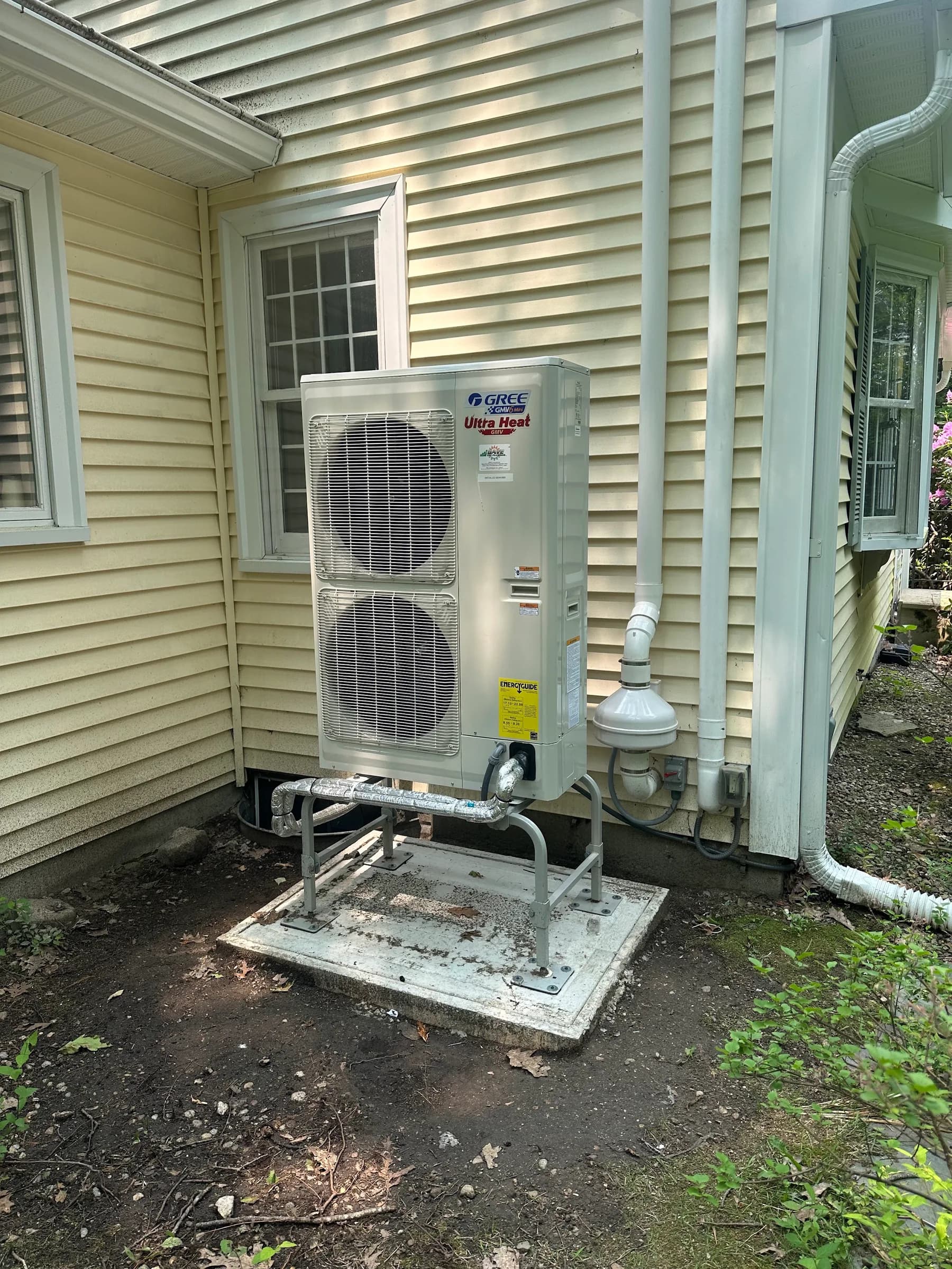 An outdoor heat pump unit on a raised stand on a concrete pad beside pale yellow siding