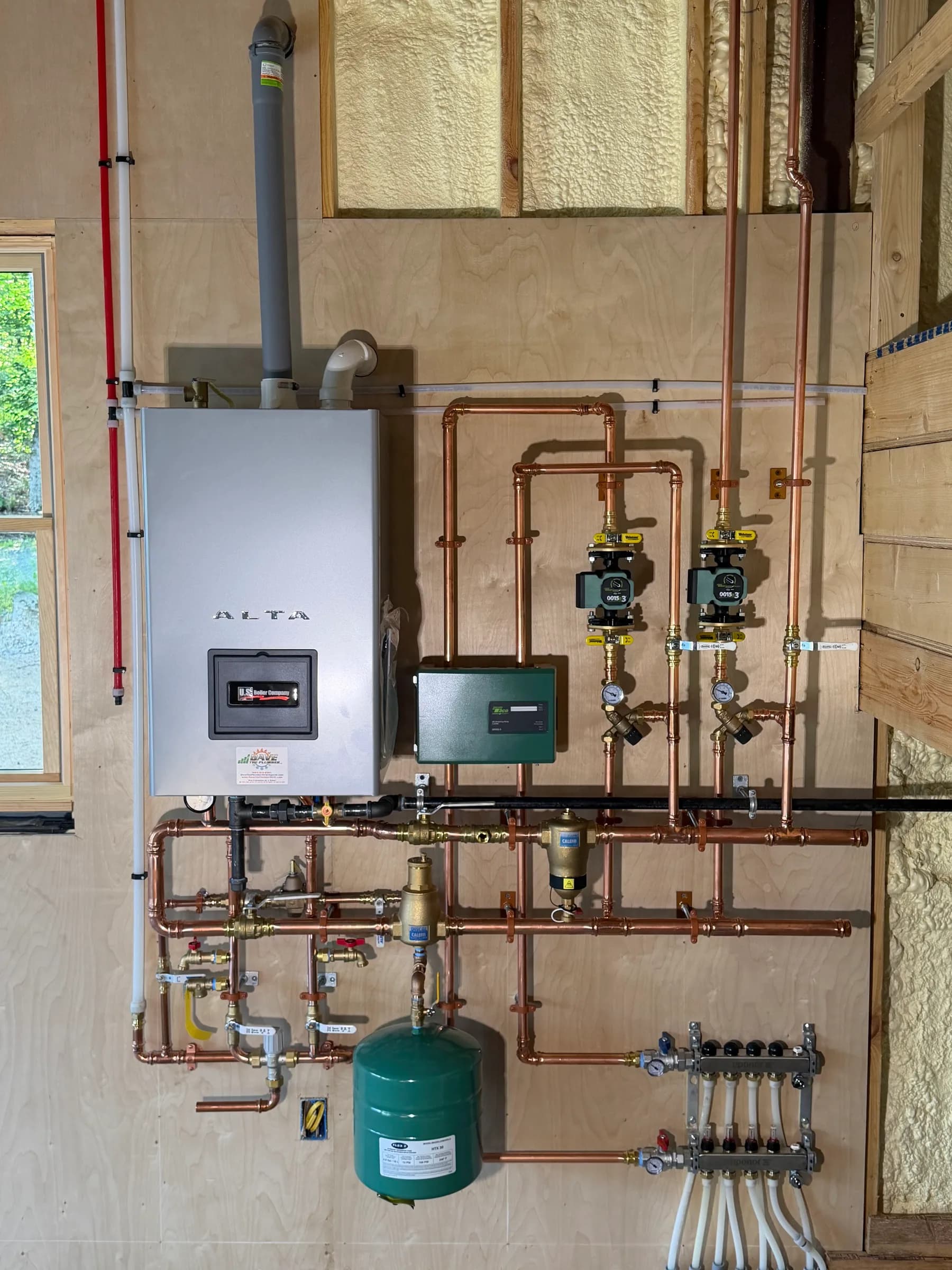 A wall hung boiler with soldered copper piping, two circulators, an expansion tank and a zone manifold in a timber framed room