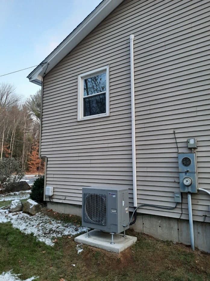 A heat pump condenser mounted on a pad beside a New Hampshire home in winter, from Dave's own job photographs