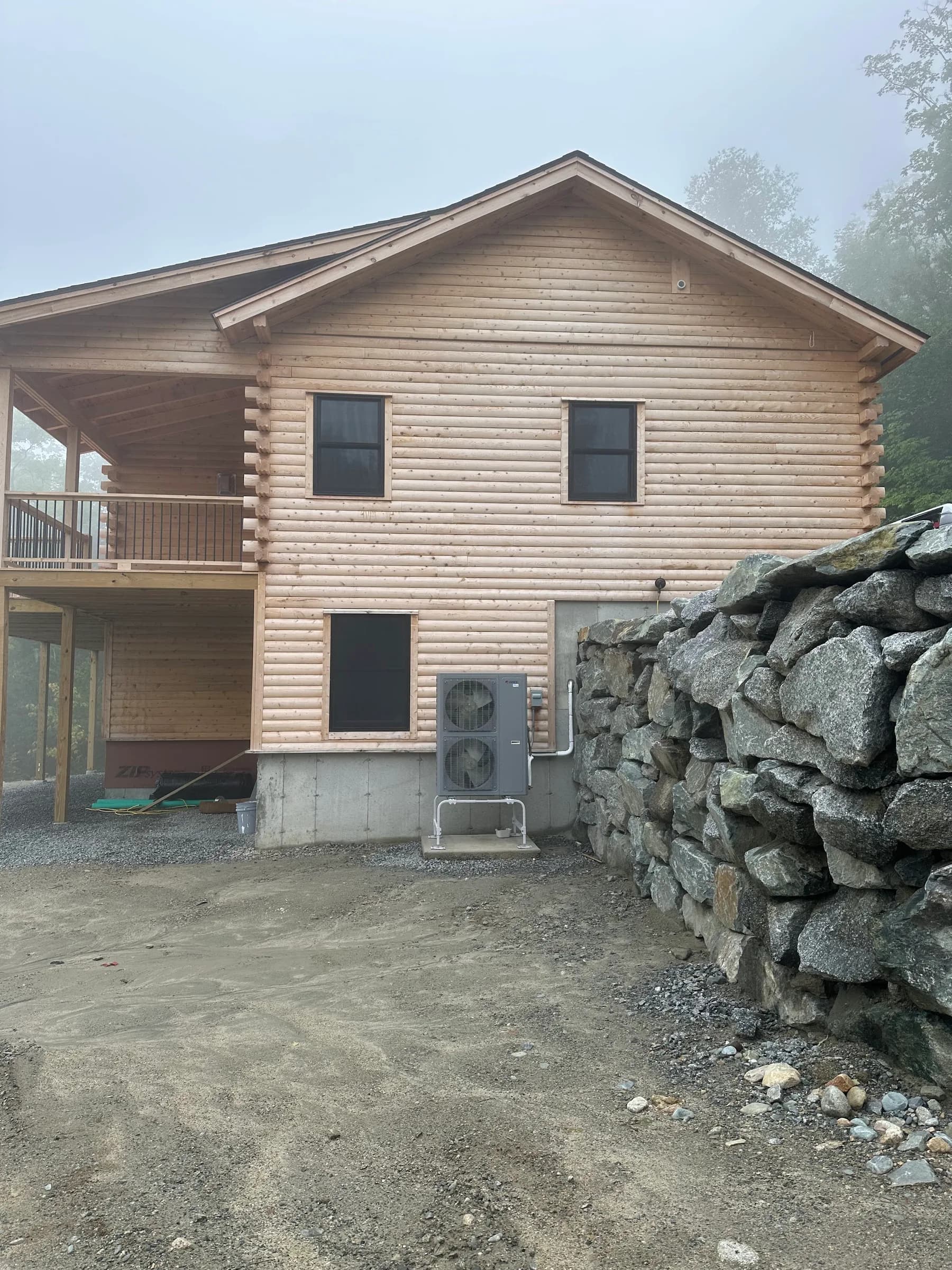 A log home in fog with a small outdoor heat pump unit at the base of the wall beside a stone wall