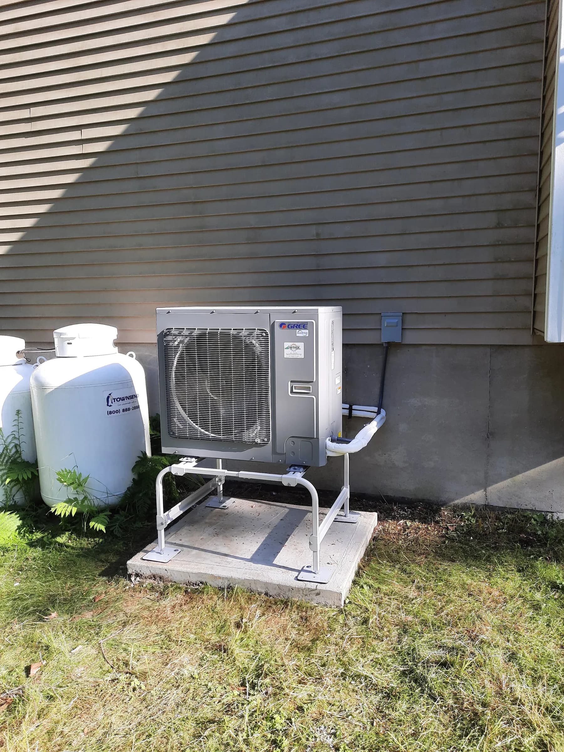 An outdoor heat pump unit on a concrete pad beside two large propane tanks at a house foundation