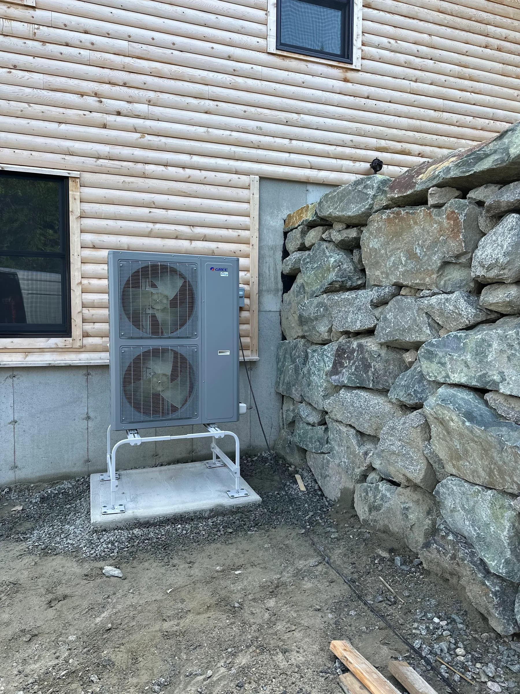 An outdoor heat pump unit on a pad beside a stone retaining wall at the corner of a timber home
