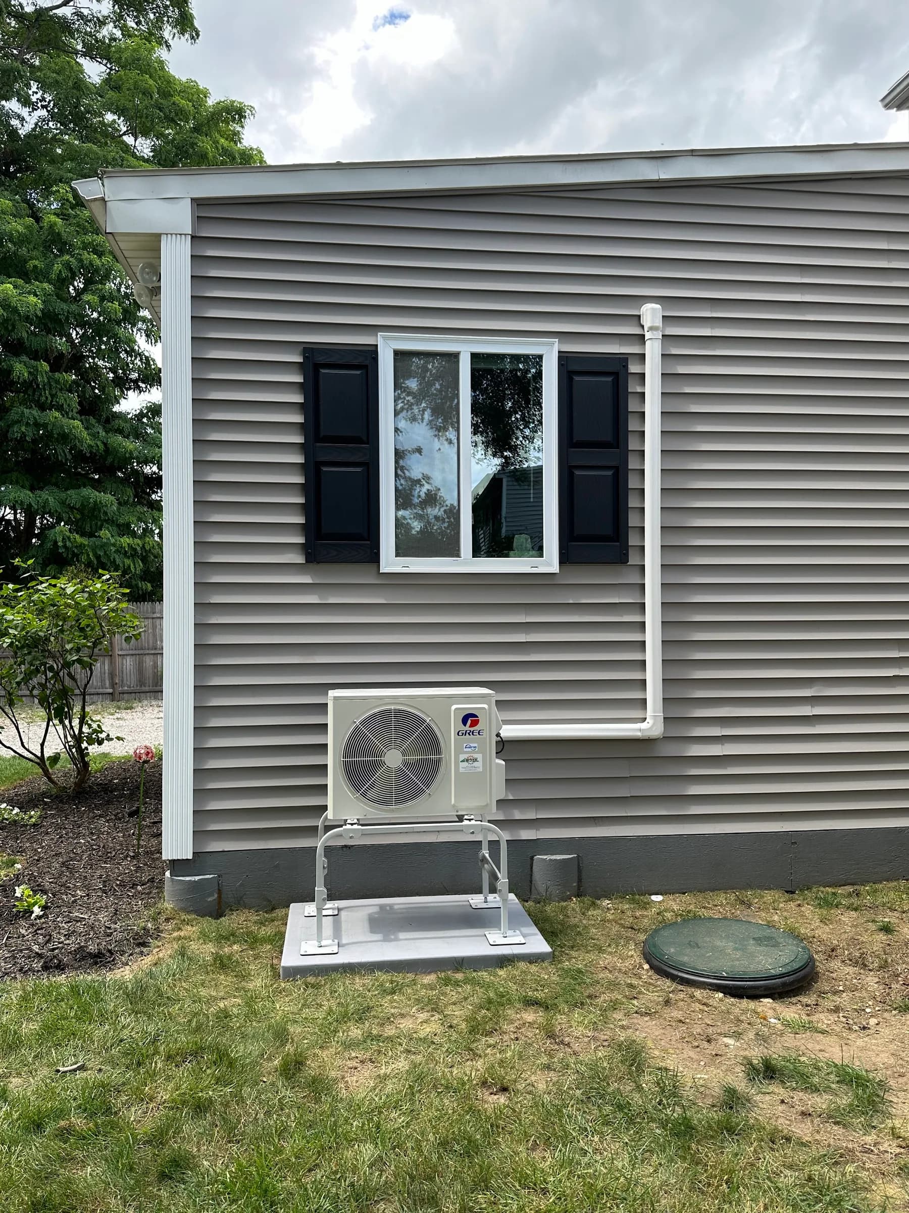 An outdoor heat pump unit on a raised stand below a window with black shutters