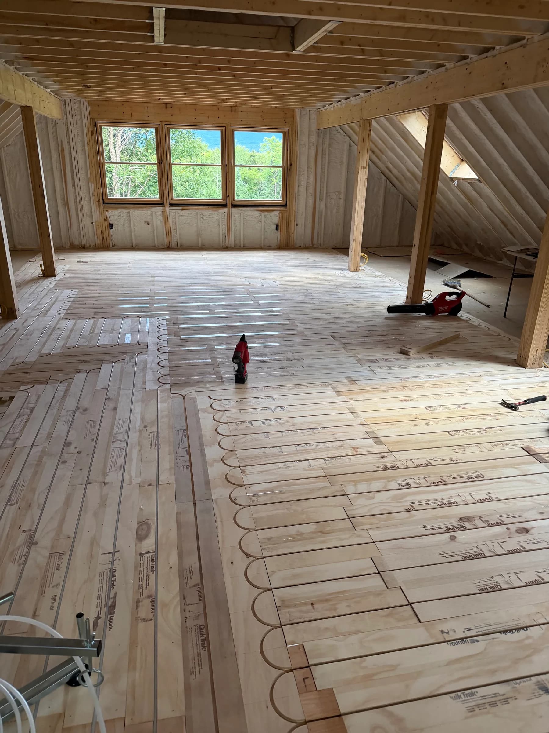 Radiant floor tubing laid over subfloor panels in a timber framed upper room with windows at the gable end