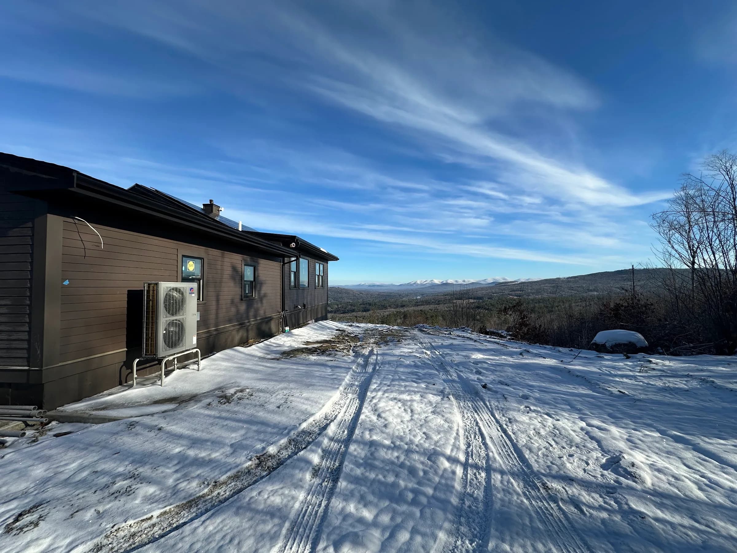Outdoor heat pump units mounted on the wall of a low house above a snow covered drive with a long ridge view behind