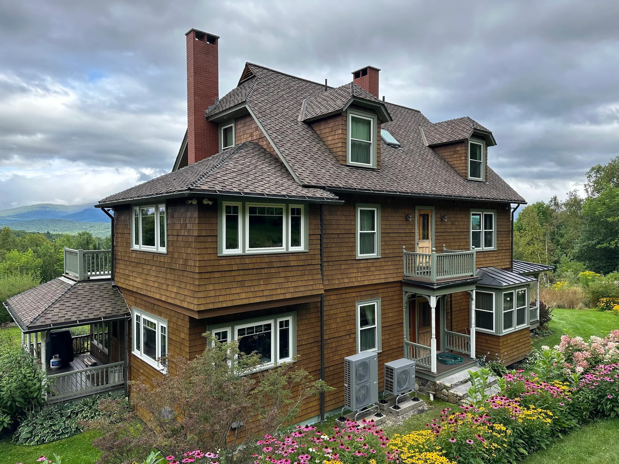 A large shingled Victorian house with heat pump units at the side elevation and planted gardens in front