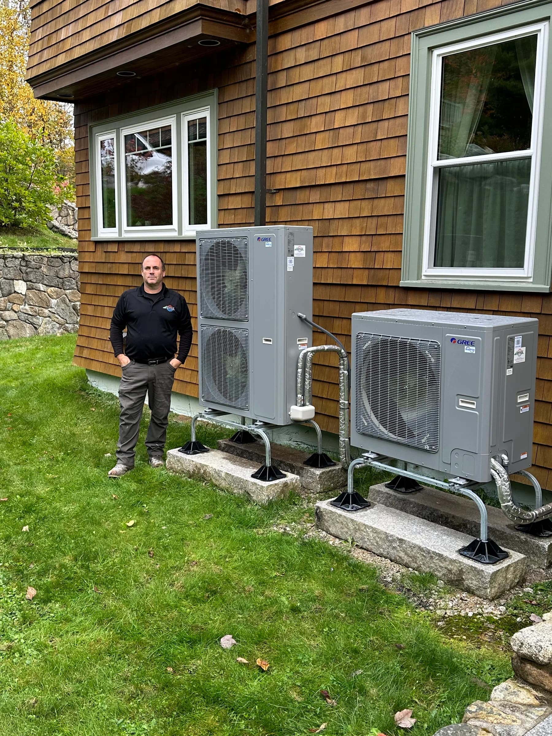 A technician standing beside two installed outdoor heat pump units at the side of a cedar shingled house