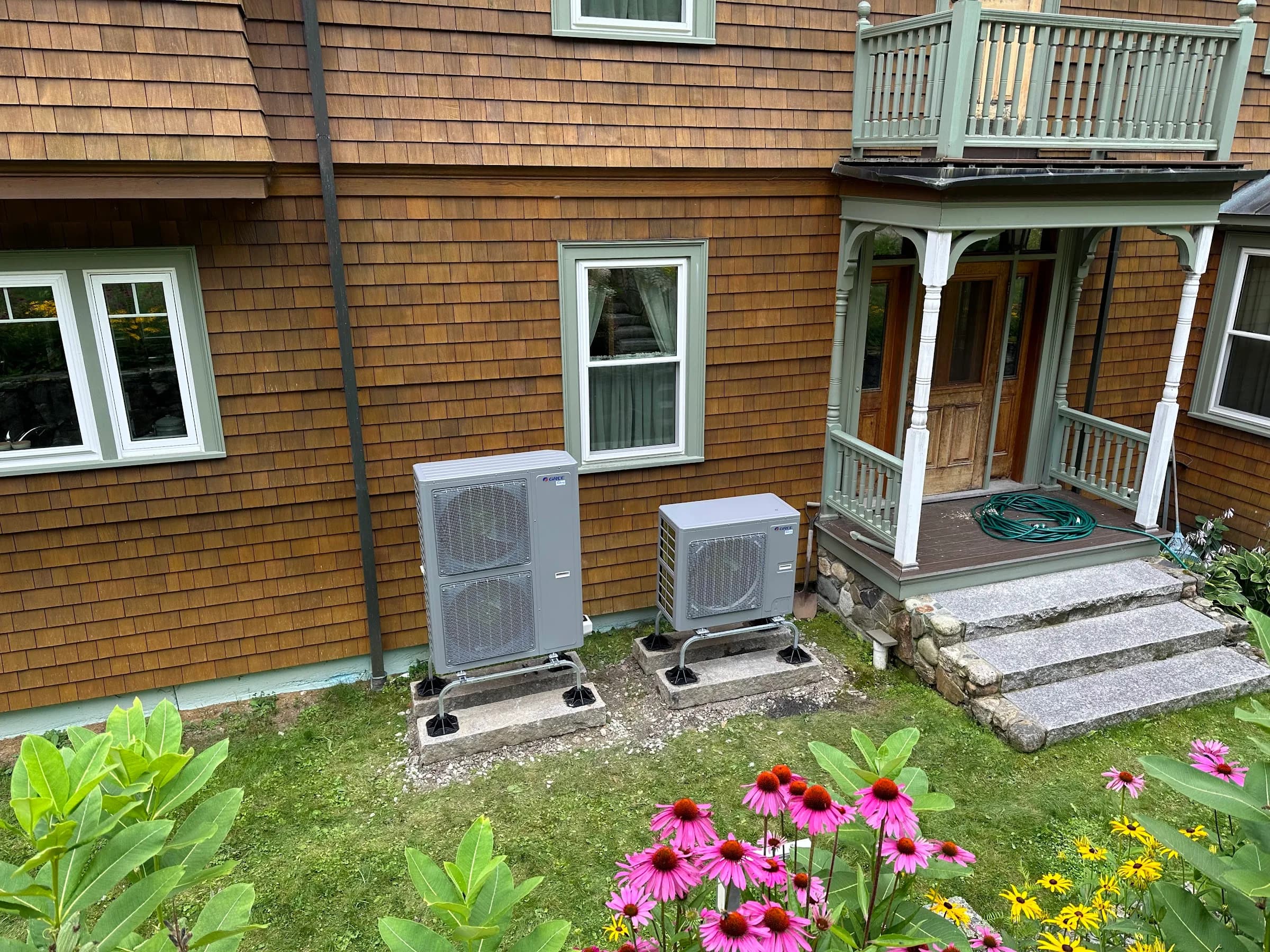 Two outdoor heat pump units beside a shingled porch with coneflowers and black eyed susans in the bed in front