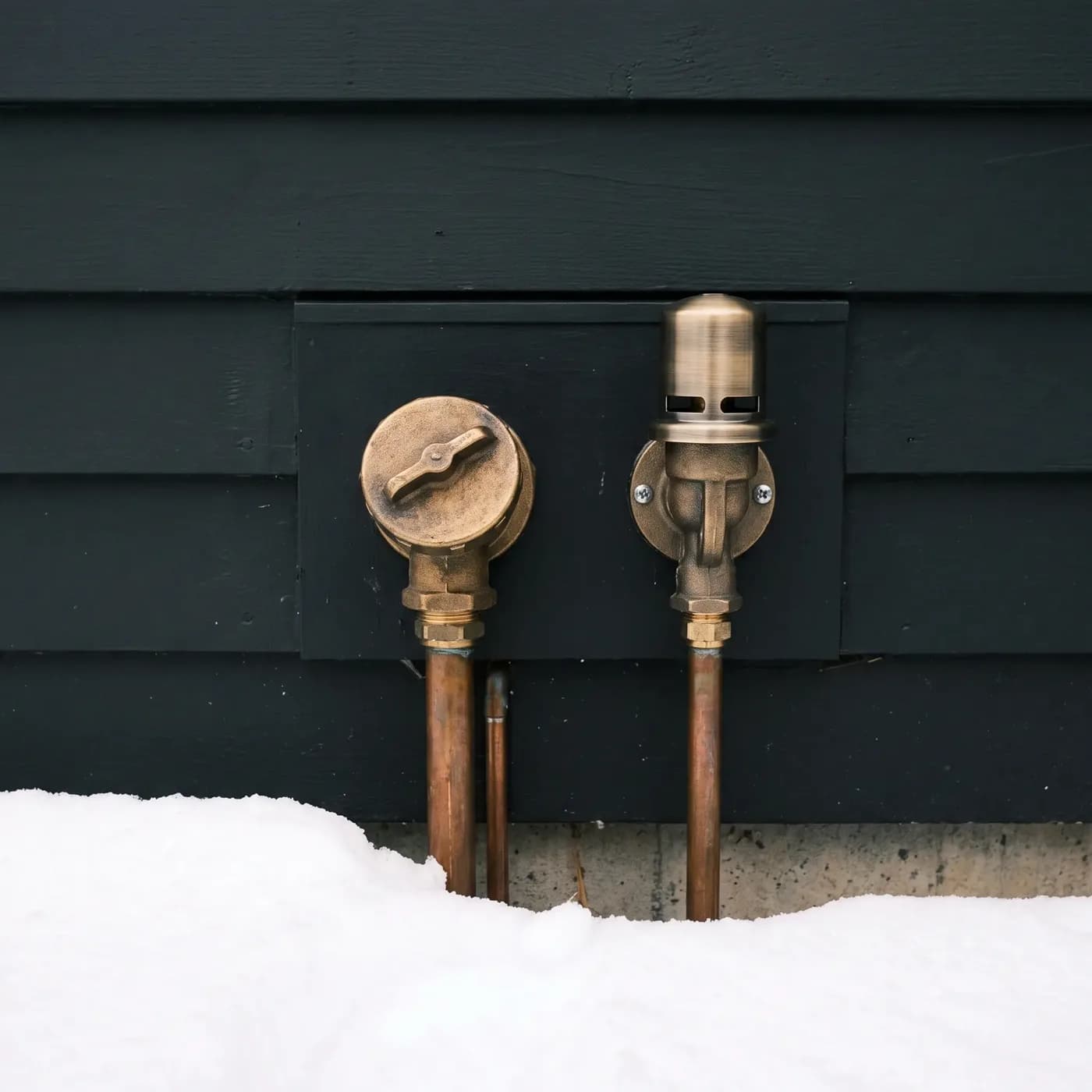 An oil fill cap and a vent alarm whistle mounted side by side on the black clapboard wall of a house, their copper lines running down into the foundation behind banked snow.