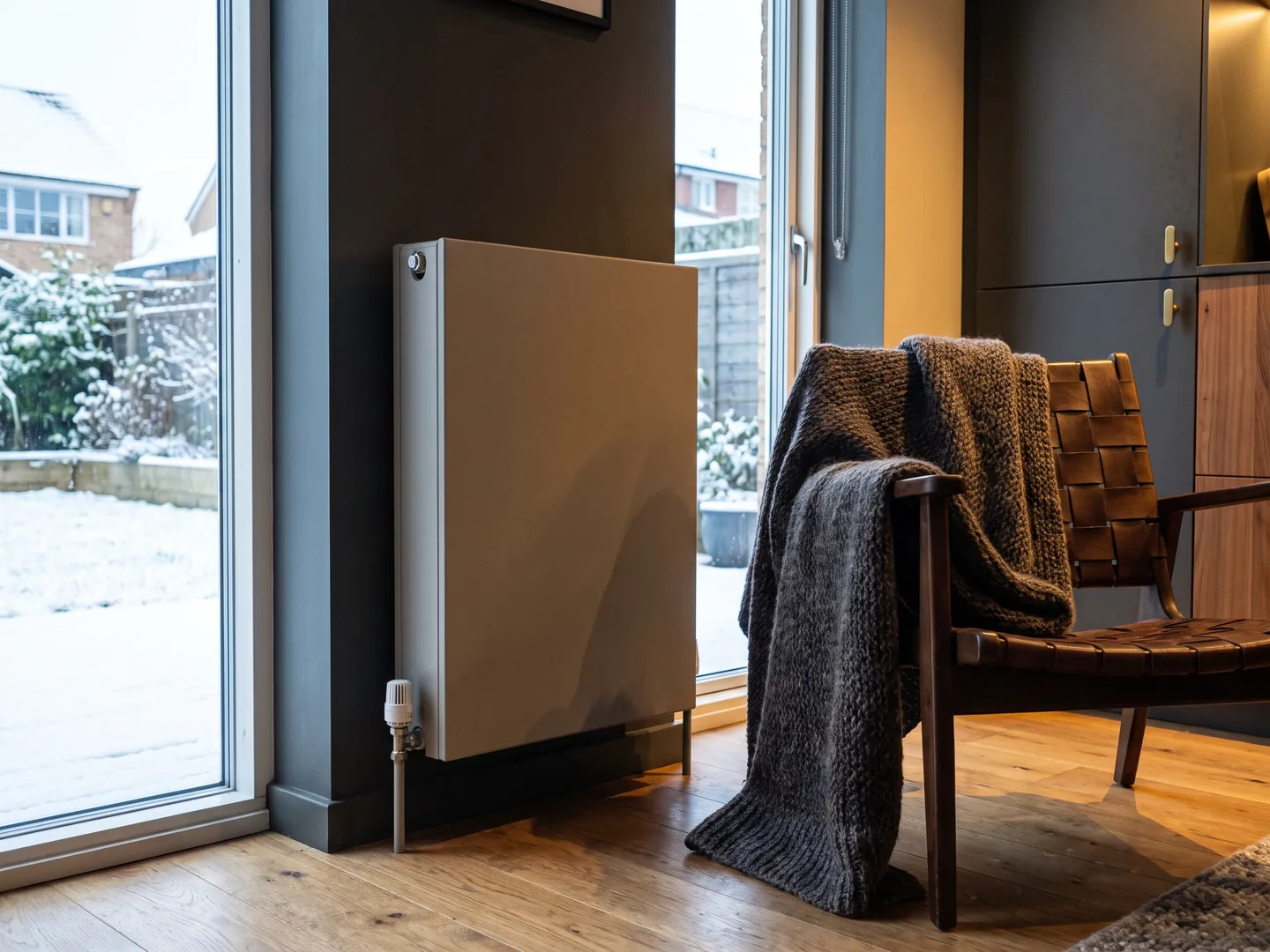 Upright panel radiator with a thermostatic valve against a charcoal wall beside a snow-covered garden